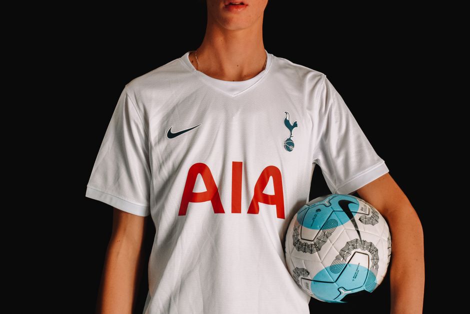 Adult soccer player in Tottenham Hotspur jersey holding a ball against a dark background.